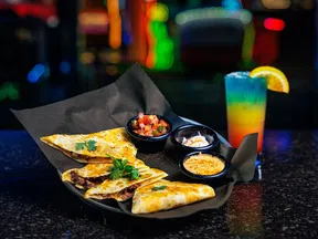 Plate of quesadillas with salsa, sour cream, and cheese dips alongside a colorful layered cocktail garnished with orange.