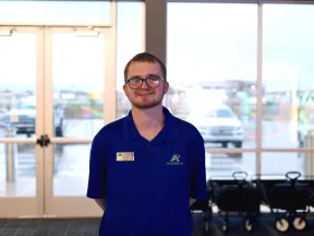 A young man in a blue polo shirt stands in a bright lobby area with large windows behind him.
