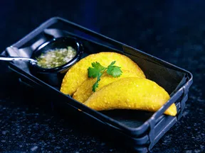 Two golden fried empanadas garnished with cilantro served in a black tray with a cup of green salsa.