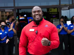 Smiling employee in red shirt welcomes guests outside, with team members in blue shirts clapping.