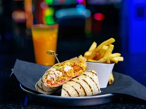 Grilled chicken wrap with fries and a drink on a plate against a dark blurred background.