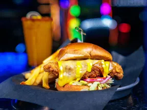 Fried chicken sandwich with melted cheese, lettuce, onions, and mustard served with fries and a drink in a colorful setting