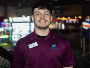 Smiling male staff member in an Andretti polo shirt, with arcade games blurred in the background.