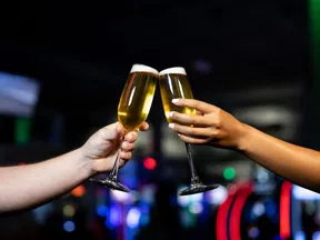 Two hands clinking champagne glasses in a festive atmosphere with colorful lights.