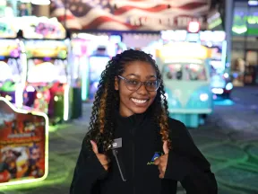 Smiling arcade employee giving thumbs up in a colorful game arcade with bright lights and machines.