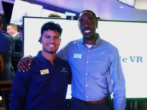 Two smiling male employees wearing name tags and company shirts pose in front of a digital display in a modern office.