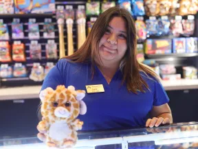 A smiling employee poses at a toy store, holding a stuffed giraffe toy, colorful merchandise in the background.
