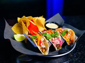 Three tacos with grilled meat, fresh vegetables, and cilantro, served with tortilla chips, lime wedge, and dipping sauce.