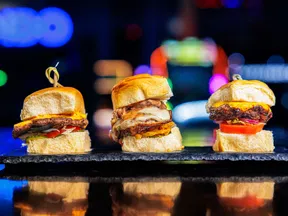Three gourmet sliders with cheese and vegetables on a black slate plate against colorful blurred background