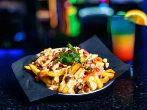 Loaded fries with cheese, ground beef, cilantro garnish, and a colorful cocktail with an orange slice on a dark table.