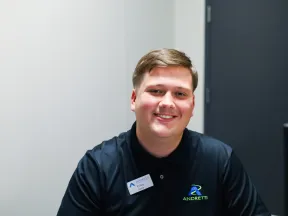 Smiling young man in a black polo shirt with Andretti logo, seated at a desk in a modern office.