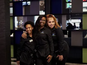 Three smiling female employees in black uniforms posing together inside a modern establishment with colorful wall panels.