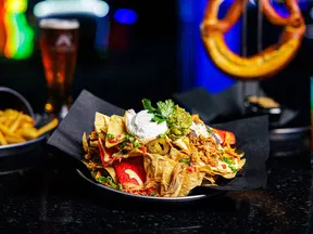 Plate of loaded nachos topped with shredded chicken, sour cream, guacamole, and jalapenos on black table in a bar.