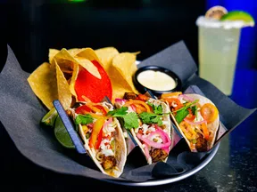 Plate of three tacos with grilled meat, pickled onions, peppers, cilantro, chips, queso dip, and a margarita glass.