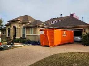 Residential house with roof under repair and large orange dumpster in driveway for construction debris removal.
