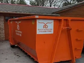Large orange dumpster with Austin Dumpsters logo and contact number parked next to a building under cloudy sky.