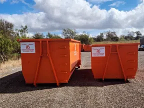 Two large orange dumpsters on gravel lot under blue sky with clouds and trees in background.