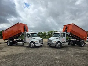 Two white dump trucks with raised orange containers parked on gravel under a cloudy sky with trees in the background