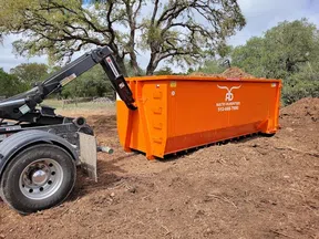 Orange Austin Dumpsters roll-off container loaded with debris on dirt ground with trees in background.