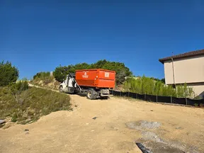 Orange dump truck on dirt road near hillside vegetation and building under clear blue sky.