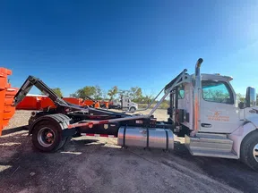 White roll-off truck with hydraulic arm parked near orange containers under clear blue sky.