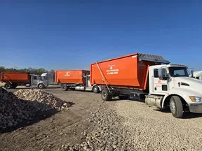 Orange dumpster trucks parked on gravel with blue sky at a construction or waste management site.