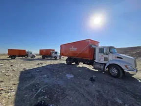 Three white trucks with large orange containers parked on barren terrain under a bright blue sky with the sun.