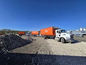 White and orange dump trucks parked on gravel near large piles of rocks under a clear blue sky.
