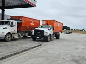 Two white trucks with orange trailers parked at a gas station on a cloudy day in a rural area