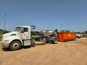 White roll-off truck attached to an orange dumpster on a dirt lot under clear blue sky