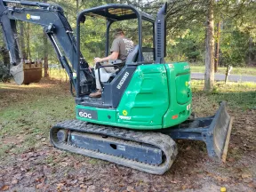 Man operating a green 60G compact excavator on a leaf-covered ground in a wooded area.