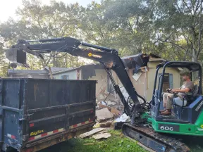 Compact excavator loading debris into a dumpster during house demolition in a wooded residential area.