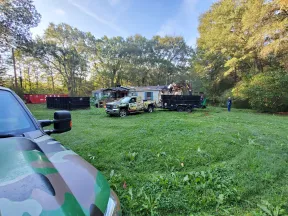 Work trucks and equipment on grassy yard with trees and a house in the background during daylight.