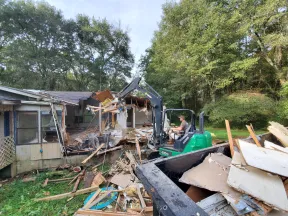 Mini excavator demolishing an old wooden house amid green trees and debris scattered on the ground.