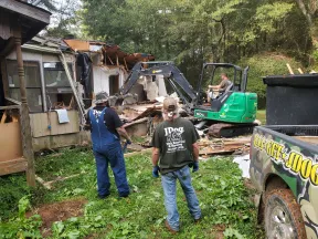 Workers using a green excavator to demolish a wooden house while two men observe from the grassy yard.