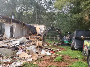 Excavator demolishing a wooden house surrounded by debris and trees in a green yard.