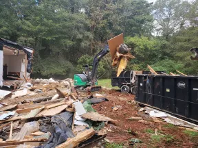 Excavator loading debris from demolished building into truck near large dumpster in wooded area.