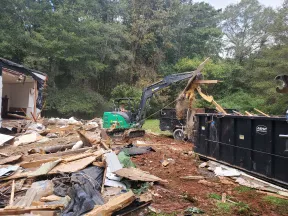 Excavator removing debris from house demolition site with dumpster in wooded area under overcast sky