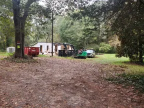 Construction site in a wooded area with excavator, pickup truck, dumpster, and demolished building in background.