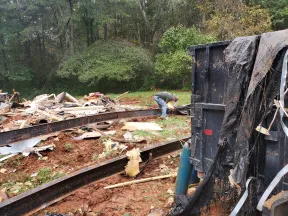 Worker welding steel beams at a construction site with scattered debris and green trees in the background.
