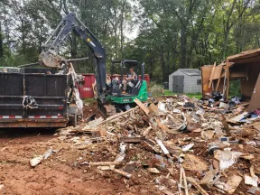 Excavator loading wooden debris and rubble into a dump truck amid a demolition site with trees and shed in background