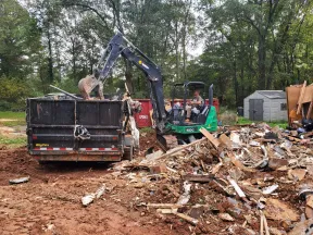 Excavator loading wooden debris into a BigTex dump trailer at a wooded demolition site.