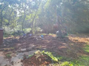 Excavator working on a cleared dirt site with debris piles and green trees under bright sunlight.