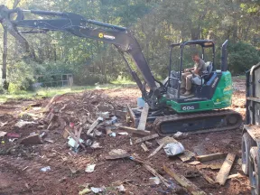 Man operating green and black mini excavator clearing wood debris in a wooded outdoor area