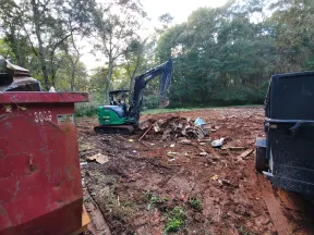 Construction site with a green excavator clearing debris and two dumpsters in a wooded area.