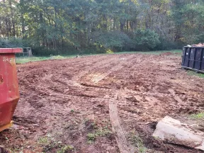 Cleared and leveled construction site with tire tracks in dirt, surrounded by trees and dumpsters.