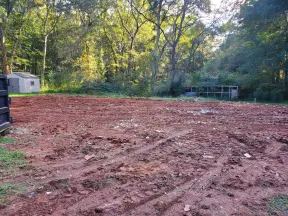 Cleared plot of red dirt land with tire tracks surrounded by trees and small outdoor sheds in the background.