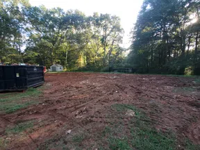 Cleared dirt land with tire tracks near green trees and black dumpsters under a bright sky.