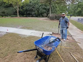 Construction worker standing beside a broken concrete sidewalk with a wheelbarrow and tools in a green yard.