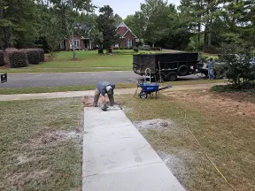 Worker smoothing wet concrete on a newly poured sidewalk in a residential neighborhood with tools and trailer nearby
